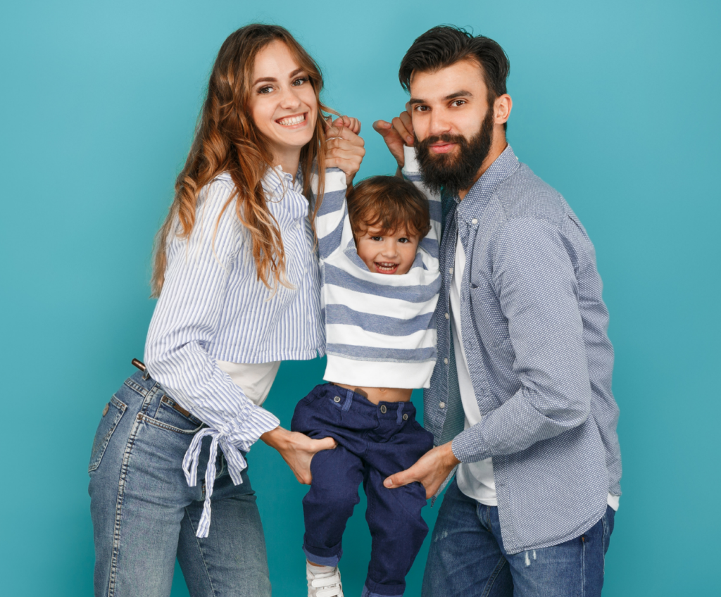 A smiling young mother and father stand together, playfully lifting their son by the hands in a joyful hug, all while looking at the camera against a blue background.