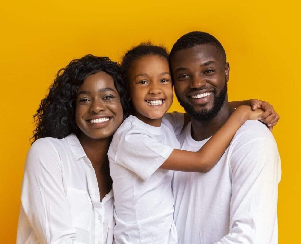 Loving family of three embracing each other and smiling while standing over yellow studio background.